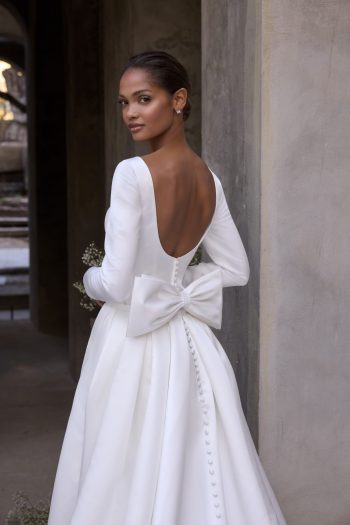 Morilee Virtue style 2965, is a satin mini wedding dress with long sleeves and a scoop low back. It also has a beautiful wrap detachable overskirt with pockets and a bow.