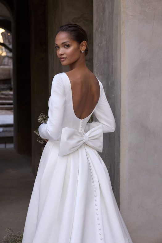 Morilee Virtue style 2965, is a satin mini wedding dress with long sleeves and a scoop low back. It also has a beautiful wrap detachable overskirt with pockets and a bow.